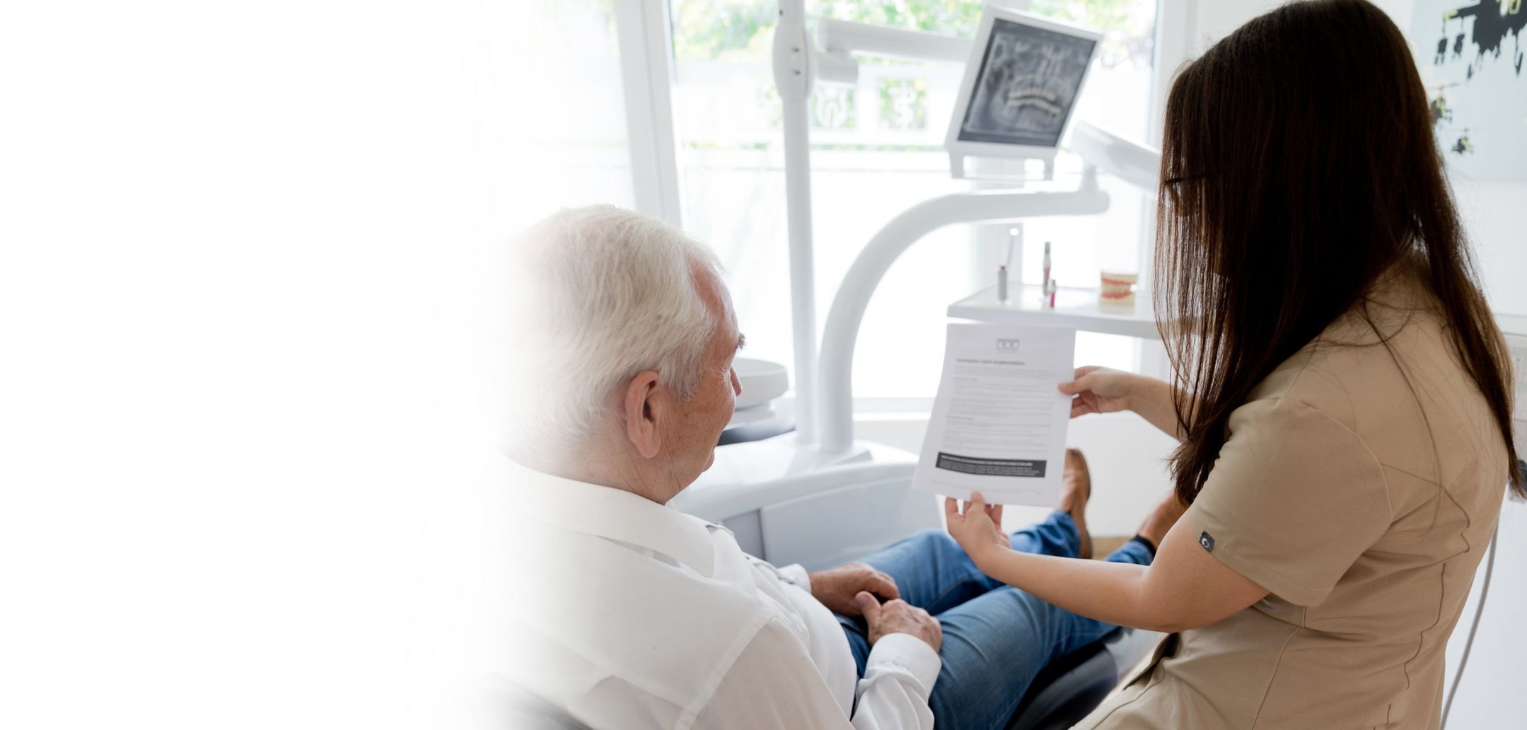 Mitarbeiterin der Praxis erläutert einem Patienten seine Behandlungsunterlagen, im Hintergrund eine Röntgenaufnahme